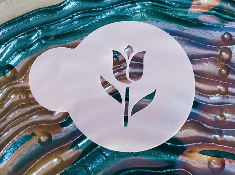 May include: A white, circular stencil featuring a tulip design. The stencil has a rounded shape with a smaller, attached section. The tulip design is cut out, revealing the background. The background has a mix of blue, green, and brown colors.