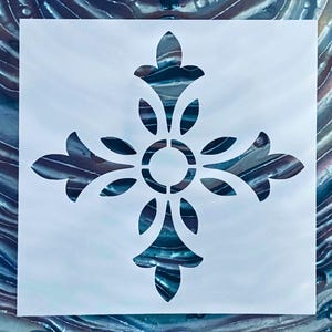 May include: A white stencil featuring a symmetrical floral cross design. The cross has four stylized floral arms with a central circular element. The background reveals a swirling blue and gray pattern, creating a layered effect.