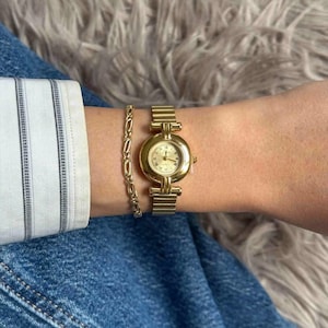 May include: A gold-toned wristwatch with a round face and a matching gold bracelet. The watch has a gold band and a light-colored watch face. The bracelet is a delicate chain design. The items are displayed on a wrist, with a blue denim background.