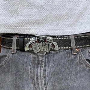 Puede incluir: Hebilla de cinturón plateada con dos revólveres y naipes. La hebilla está sujeta a un cinturón de cuero negro con tachuelas decorativas y motivos florales. El cinturón se lleva con vaqueros grises.