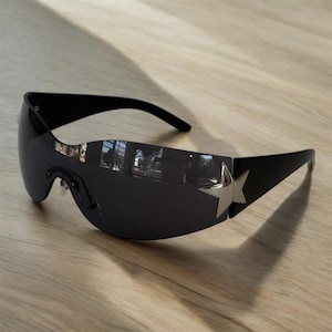 Puede incluir: Gafas de sol negras con diseño envolvente y un adorno de estrella plateada en las patillas. Las lentes son oscuras y las monturas son elegantes y modernas, sobre una superficie de madera clara.