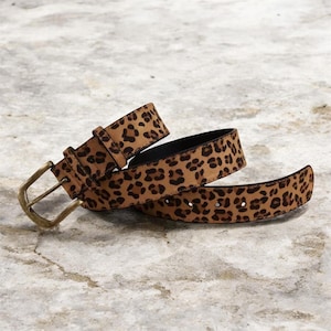 Puede incluir: Un cinturón marrón con estampado de leopardo. El cinturón tiene una hebilla rectangular de color dorado y una parte inferior negra. El cinturón está hecho de un material suave y se muestra enrollado sobre una superficie texturizada.