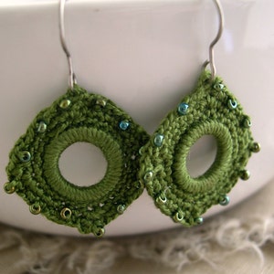 May include: A pair of handmade green crochet earrings with a circular center and beaded accents. The earrings are diamond-shaped with a hook for pierced ears. The beads are a mix of green and blue.