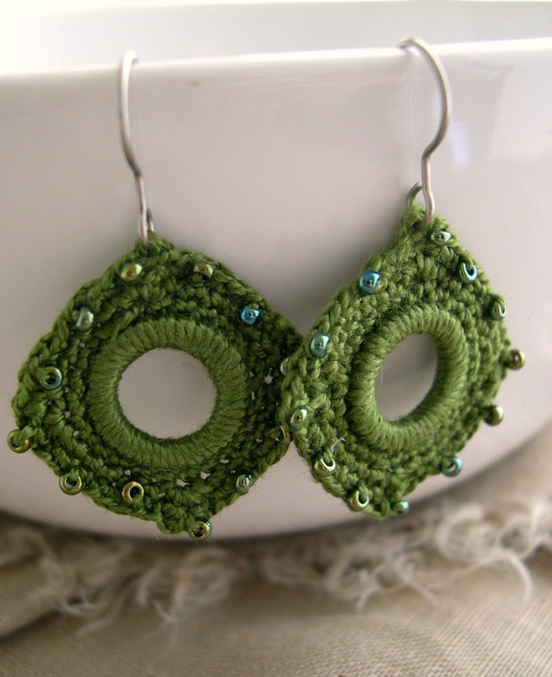 May include: A pair of handmade green crochet earrings with a circular center and beaded accents. The earrings are diamond-shaped with a hook for pierced ears. The beads are a mix of green and blue.