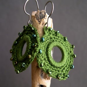 May include: A pair of handmade green crochet earrings with a circular design and a central hole. The earrings are adorned with small, iridescent beads and suspended from silver-colored ear wires. The earrings are displayed on a piece of driftwood.