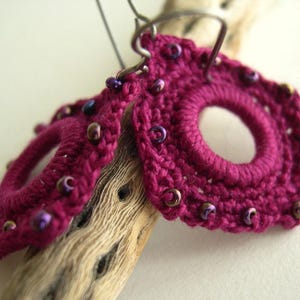 May include: A pair of handmade magenta crochet earrings with a circular design and a central opening. The earrings are adorned with small, iridescent beads and are suspended from silver-tone ear wires. The earrings are displayed on a piece of driftwood.
