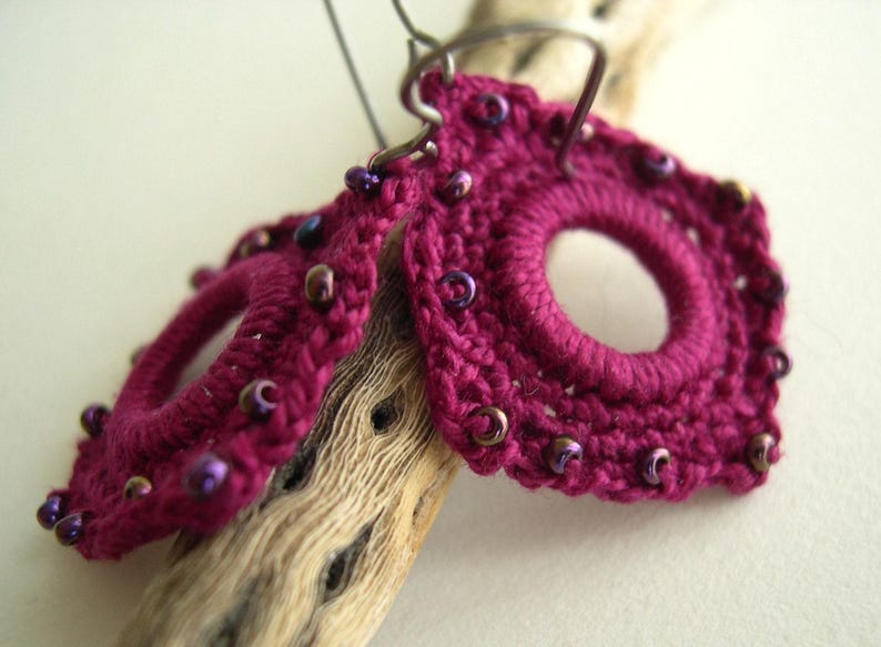 May include: A pair of handmade magenta crochet earrings with a circular design and a central opening. The earrings are adorned with small, iridescent beads and are suspended from silver-tone ear wires. The earrings are displayed on a piece of driftwood.