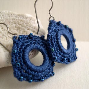 May include: A pair of handmade, dark blue crochet earrings. Each earring is circular with a central hole and is adorned with small, blue beads around the edge. The earrings hang from silver-colored ear wires.