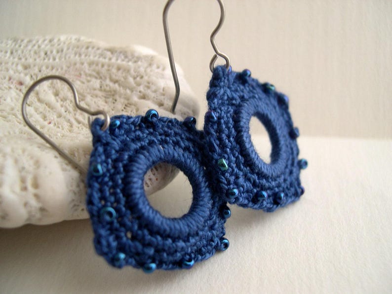 May include: A pair of handmade, dark blue crochet earrings. Each earring is circular with a central hole and is adorned with small, blue beads around the edge. The earrings hang from silver-colored ear wires.