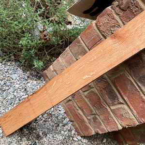 May include: A close-up of a long, narrow piece of wood with a reddish-brown color. The wood has a smooth surface with some visible grain patterns. The wood is lying on a bed of small, gray stones.