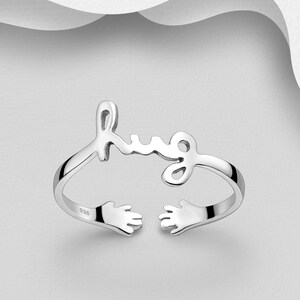 May include: Silver ring with the word "hug" written in cursive and two hands on either side of the word.