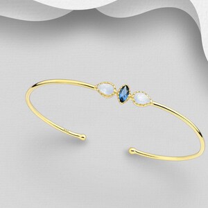 May include: A gold bangle bracelet with three oval-shaped stones. The center stone is a dark blue gemstone, and the two outer stones are white.