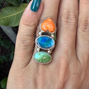 Pode incluir: Um anel de prata esterlina com três pedras empilhadas verticalmente: uma pedra laranja em forma de coração no topo, uma opala azul iridescente redonda no centro e uma pedra turquesa oval verde na base, engastadas em molduras de metal serrilhadas.