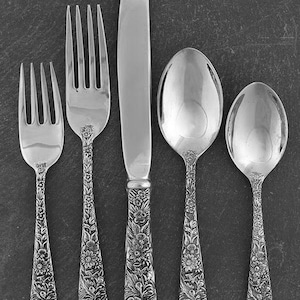 May include: A five-piece silver cutlery set comprising a salad fork, a dinner fork, a dinner knife, a soup spoon, and a teaspoon. Each item features a detailed, embossed floral pattern along the handle, with polished, smooth metal bowls and blades.