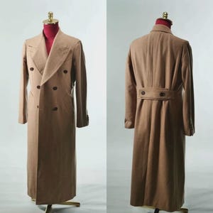 May include: A long, double-breasted tan coat featuring wide lapels, dark brown buttons on the front, and a matching buttoned belt detail across the back. The coat is displayed on a mannequin, showing both the front and back views of the garment.
