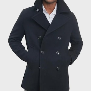 May include: A dark navy blue double-breasted pea coat featuring a wide notched lapel, a high collar, and six black buttons arranged in two vertical rows. The coat is worn over a white collared shirt and paired with grey plaid trousers.