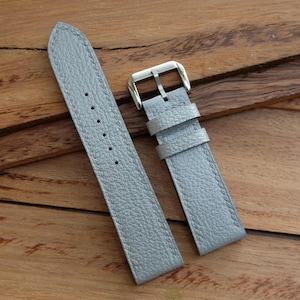 May include: A light gray leather watch strap with a silver-colored buckle. The strap has a textured surface and is stitched with a matching gray thread. The watch band is shown on a wooden surface.