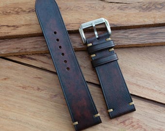 May include: A dark brown leather watch strap with a silver buckle. The strap has a distressed finish with red undertones and is stitched with light tan thread. The watch band is shown on a wooden surface.