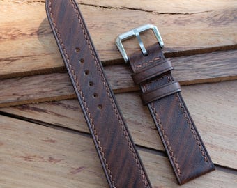 May include: A brown leather watch strap with a silver buckle. The strap has a textured surface and is stitched with a lighter brown thread. The watch band is displayed on a wooden surface.
