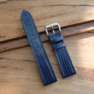May include: A pair of navy blue watch straps with a textured, reptile-like pattern. One strap has a silver-colored buckle. The straps are displayed on a wooden surface.