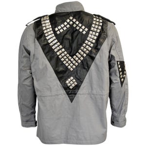May include: A gray jacket with a black leather panel on the back, adorned with silver-colored square studs. The jacket has a collar, shoulder epaulets, and a pocket on the left sleeve. The back panel is shaped in a V-design.