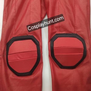 Pu&ograve; includere: Primo piano di pantaloni rossi di un costume con dettagli neri intorno alle ginocchiere. I pantaloni hanno un motivo strutturato e il testo "Cosplayhunt.com" &egrave; visibile.