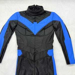 Può includere: Un costume da supereroe nero e blu con collo alto e maniche lunghe. Il costume presenta accenti blu sulle spalle, sulle braccia e sul petto, con un'emblema a forma di pipistrello sul petto. La tuta è realizzata in un materiale strutturato.