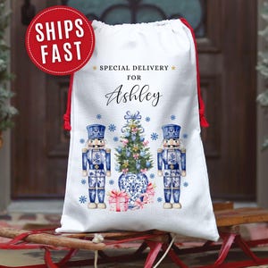 May include: White Christmas gift bag with red drawstring closure. Features a blue and white nutcracker design, a Christmas tree, and the text "Special Delivery for Ashley". A red circle with "SHIPS FAST" is in the upper left.