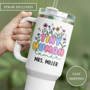 May include: White insulated tumbler with a handle, featuring the text "Tiny Human Tamer" in colorful bubble letters, surrounded by floral illustrations. The tumbler includes a straw and the name "Mrs. Miller".