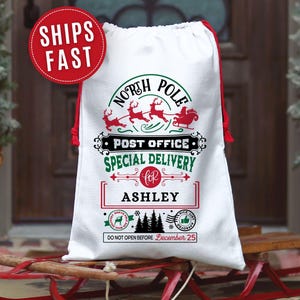 May include: White Christmas gift bag with red drawstring and festive graphics. The bag features the text "North Pole Post Office Special Delivery" with reindeer and a sleigh. The name "Ashley" is printed below, with the text "Do Not Open Before December 25".