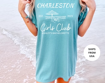 May include: Seafoam green t-shirt with white text that reads "Charleston Girls Club Ashley's Bachelorette" with a beach umbrella and chair graphic. The shirt is a relaxed fit.