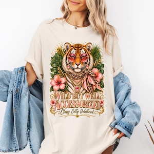 Könnte beinhalten: Ein cremefarbenes T-Shirt mit einer Grafik eines Tigers, der eine rosa Sonnenbrille, einen Schal mit Leopardenmuster und goldenen Schmuck trägt. Das Design enthält tropische Hibiskusblüten und grüne Blätter mit dem Text WILD BUT WELL ACCESSORIZED Classy. Catty. Unbothered.