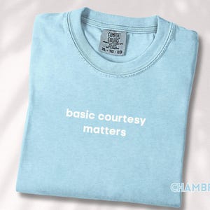 May include: A light blue t-shirt with the words "basic courtesy matters" printed in white. The shirt is folded and has a tag that says "Comfort Colors". The word "CHAMBRAY" is printed on the bottom right corner.
