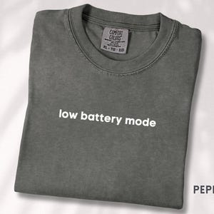 May include: Pepper-colored t-shirt with the phrase "low battery mode" in white text. The shirt is folded and has a Comfort Colors tag. The word "PEPPER" is printed in the bottom right corner.