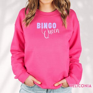 May include: A bright pink long-sleeve crewneck sweatshirt featuring the text BINGO in light purple block letters and the word Queen in white cursive script centered on the chest. The garment has a ribbed neckline, cuffs, and hem.