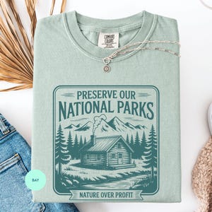 May include: A sage green short-sleeve t-shirt featuring a dark teal graphic of a cabin in the woods with mountains and trees. The text reads PRESERVE OUR NATIONAL PARKS and NATURE OVER PROFIT. A silver chain necklace rests on the collar area.