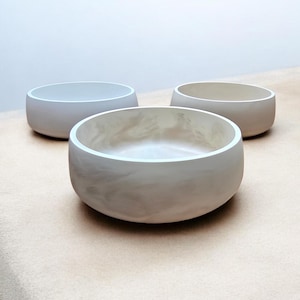 May include: Three round, light-colored bowls with a smooth, matte finish. The bowls are arranged in a group, with one in the foreground and two in the background. The interior of the bowls has a marbled effect.