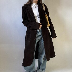 May include: A long, dark brown corduroy coat with a button-up front and two front pockets. The coat is worn over a white top and blue jeans. A tan tote bag is carried over the shoulder.