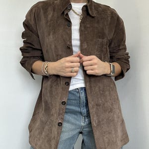 May include: A brown suede shirt with a button-down front and a single chest pocket. The shirt is worn over a white t-shirt and paired with blue jeans. The sleeves are long and buttoned at the cuffs. The shirt has a relaxed fit.