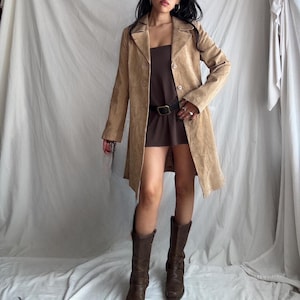 May include: A tan suede coat with a button closure, worn over a brown dress and black belt. The outfit is completed with brown knee-high boots. The coat has a classic lapel and a tailored fit, suitable for a vintage or bohemian style.