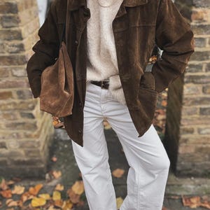 May include: A brown suede jacket with a collared neckline and button closure. The jacket is paired with a cream-colored sweater, white pants, and a brown suede tote bag. The outfit is completed with a brown belt and black shoes.