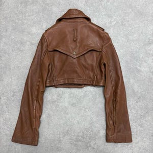 May include: A brown leather jacket with a cropped design. The jacket features a notched collar, button details, and long sleeves. The back of the jacket is visible, showcasing the tailored construction and a small button closure.