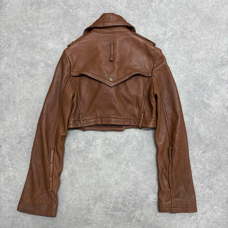 May include: A brown leather jacket with a cropped design. The jacket features a notched collar, button details, and long sleeves. The back of the jacket is visible, showcasing the tailored construction and a small button closure.