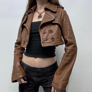 May include: A cropped brown leather jacket with a double-breasted design. The jacket features a notched collar, button closures, and long sleeves. The jacket is worn over a black top and paired with dark-wash jeans.