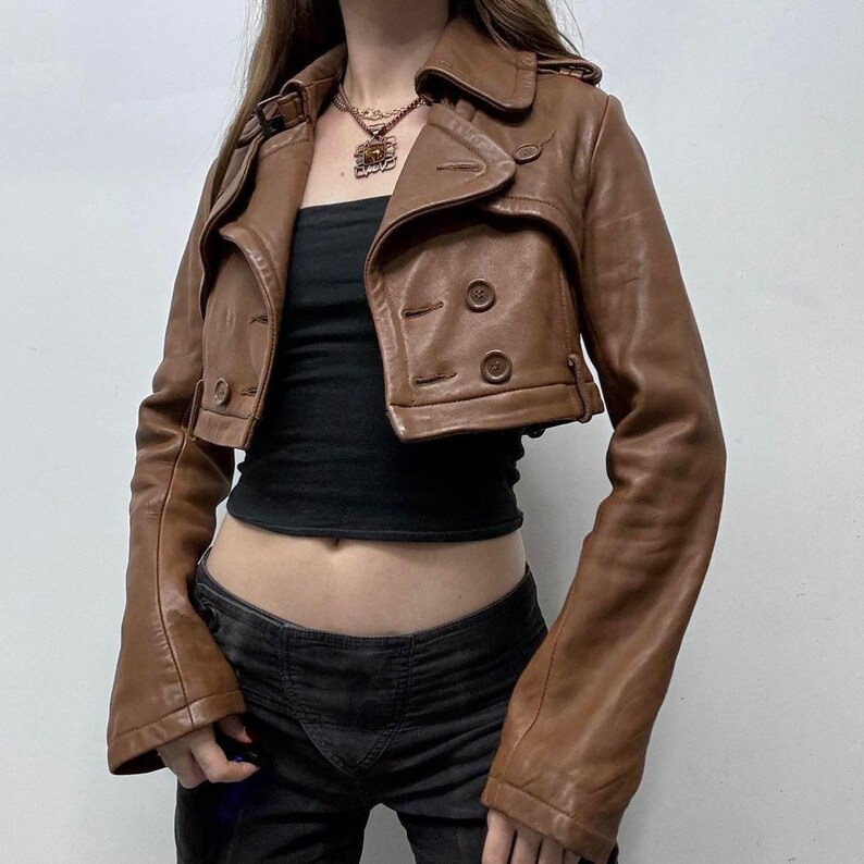 May include: A cropped brown leather jacket with a double-breasted design. The jacket features a notched collar, button closures, and long sleeves. The jacket is worn over a black top and paired with dark-wash jeans.