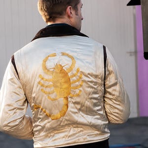 May include: A cream-colored quilted satin bomber jacket featuring a large, embroidered gold scorpion on the back. The jacket has a dark brown collar and dark brown ribbed cuffs at the waist. The fabric has a subtle sheen and diamond-pattern stitching.