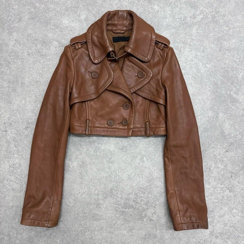 May include: A brown leather jacket with a cropped design. The jacket features a double-breasted closure with four buttons, a wide collar, and long sleeves. The jacket is a warm brown color and appears to be made of genuine leather.