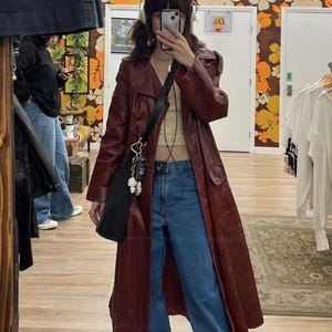 May include: A woman wearing a long, burgundy leather trench coat with a black bag and a silver chain necklace. She is also wearing blue jeans and a white crop top.