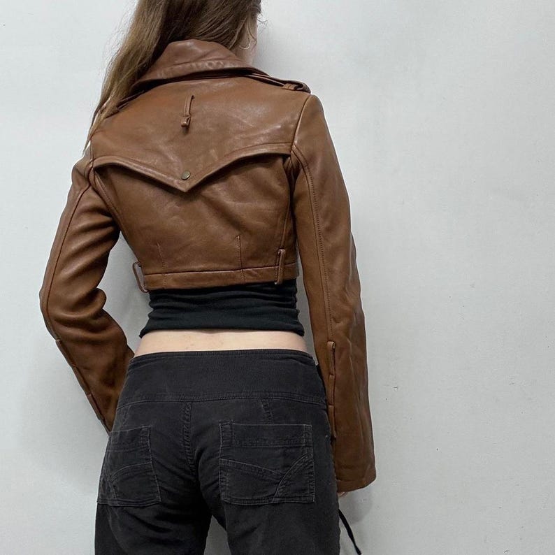 May include: A brown leather jacket with a cropped design. The jacket features a notched collar, long sleeves, and a button closure. The back view shows the jacket's tailored fit and details.