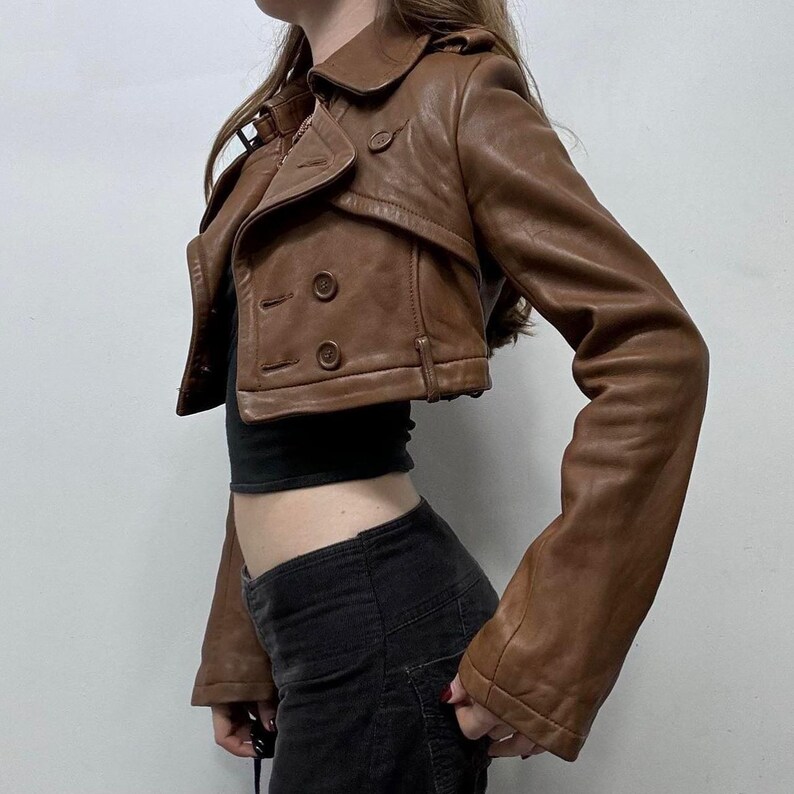 May include: A brown leather cropped jacket with a double-breasted design. The jacket features a notched collar, two rows of buttons, and a fitted silhouette. The jacket's color is a warm, medium brown.